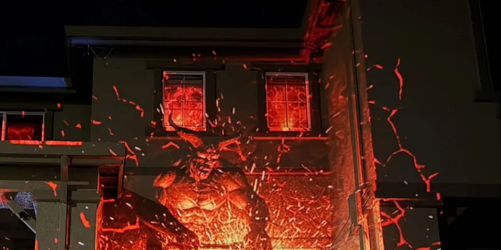 Projection of a horned demon hammering a weapon on a fiery anvil, sparks flying with glowing orange and red accents across the house exterior.
