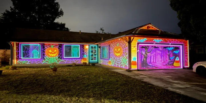 House covered in glowing Halloween projections featuring spiderwebs, jack-o-lanterns, bats, and a spooky purple forest on the garage door.